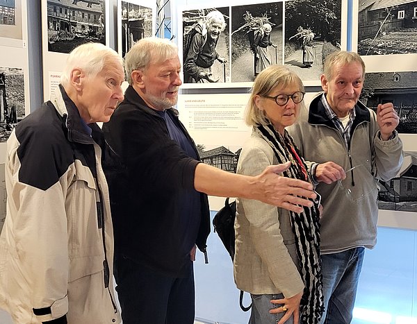 Drei Männer und eine Frau stehen an Fotowänden in einem Gebäude. Einer der Männer deutet mit seiner rechten Hand auf eine Foto. Die anderen schauen interessiert dorthin.